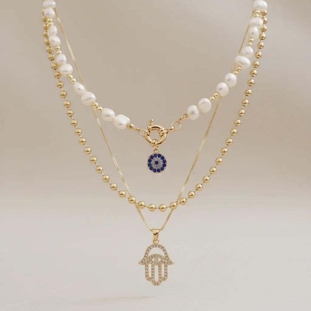 Composition de colliers multi-rangs style bohème chic : choker perles d'eau douce avec pendentif mauvais œil bleu, chaîne à billes dorée et collier main de Fatma strass.