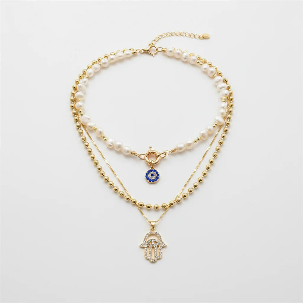 Composition de colliers multi-rangs style bohème chic : choker perles d'eau douce avec pendentif mauvais œil bleu, chaîne à billes dorée et collier main de Fatma strass.