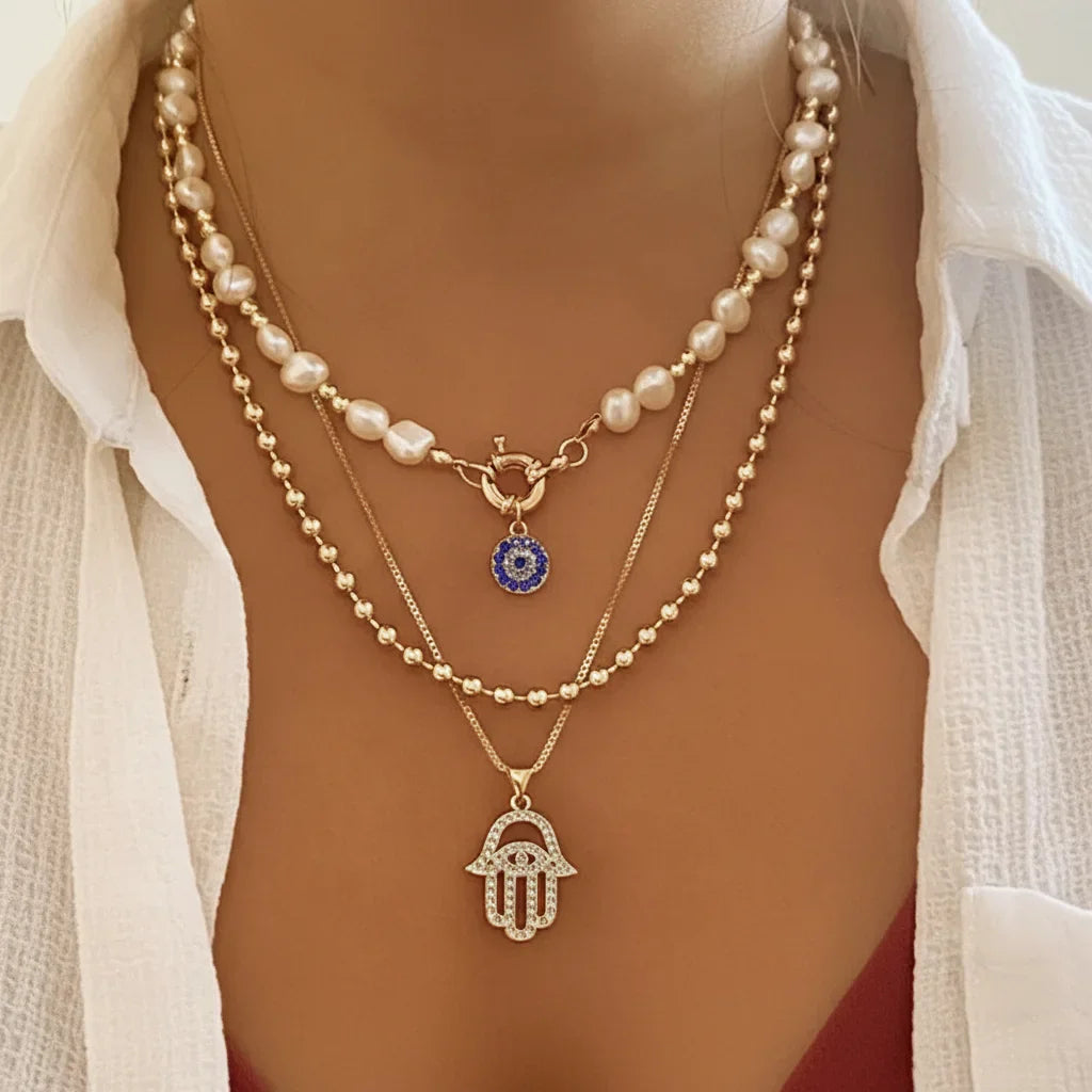 Composition de colliers multi-rangs style bohème chic : choker perles d'eau douce avec pendentif mauvais œil bleu, chaîne à billes dorée et collier main de Fatma strass.